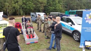 Kobieta żołnierz siedzi na symulatorze wypadku. Obok niej stoją inni żołnierze oraz osoby cywilne czekające w kolejce. Na stoisku promocyjnym policji znajduje się także radiowóz.