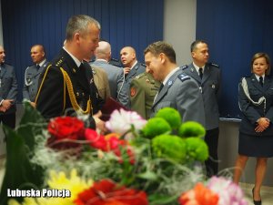 Na pierwszym planie bukiet kwiatów. Na drugim planie Młodszy Inspektor Mirosław Kędziora przyjmuje gratulacje z okazji objęcia stanowiska Komendanta od Komendanta Powiatowej Straży Pożarnej.